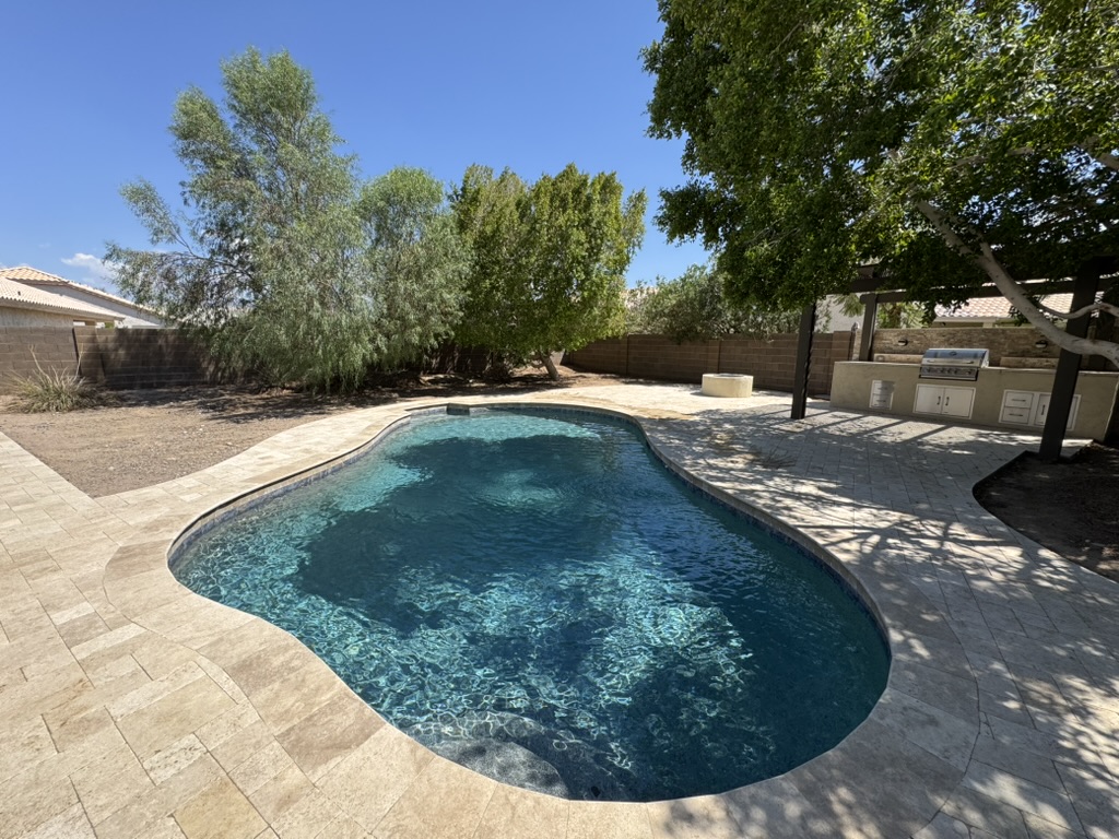 A freeform backyard pool surrounded by a stone patio and mature trees